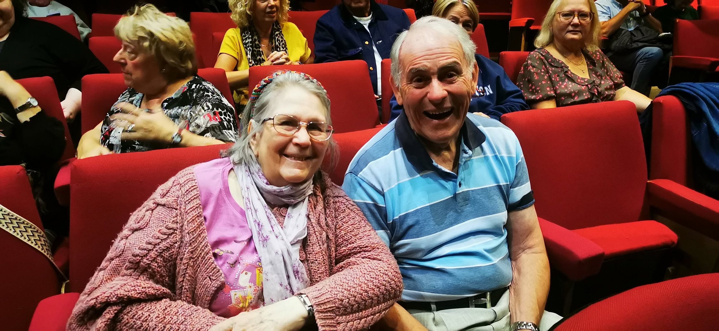 Two elderly individuals, a woman with glasses and a man in a striped polo shirt, sitting in red theater seats, smiling at the camera. Several other people are seated around them, engaged in conversations or looking elsewhere in a theater or auditoriu