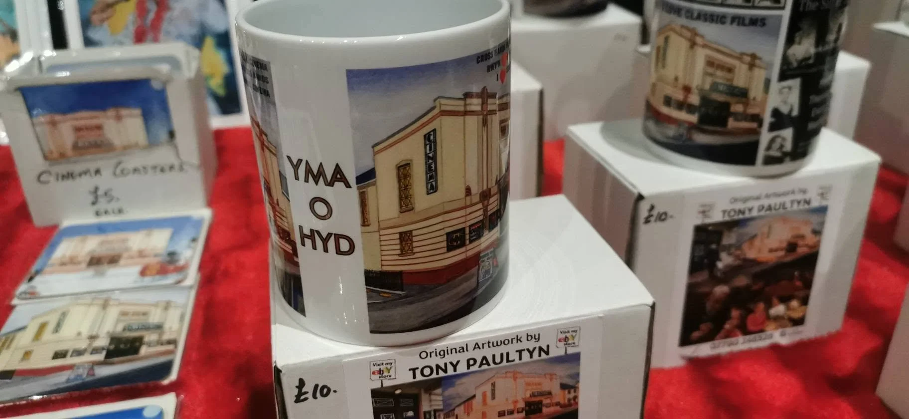 A white ceramic mug featuring artwork of a building on a boxed stand, with other similar mugs and boxes in the background, displayed on a red cloth table.