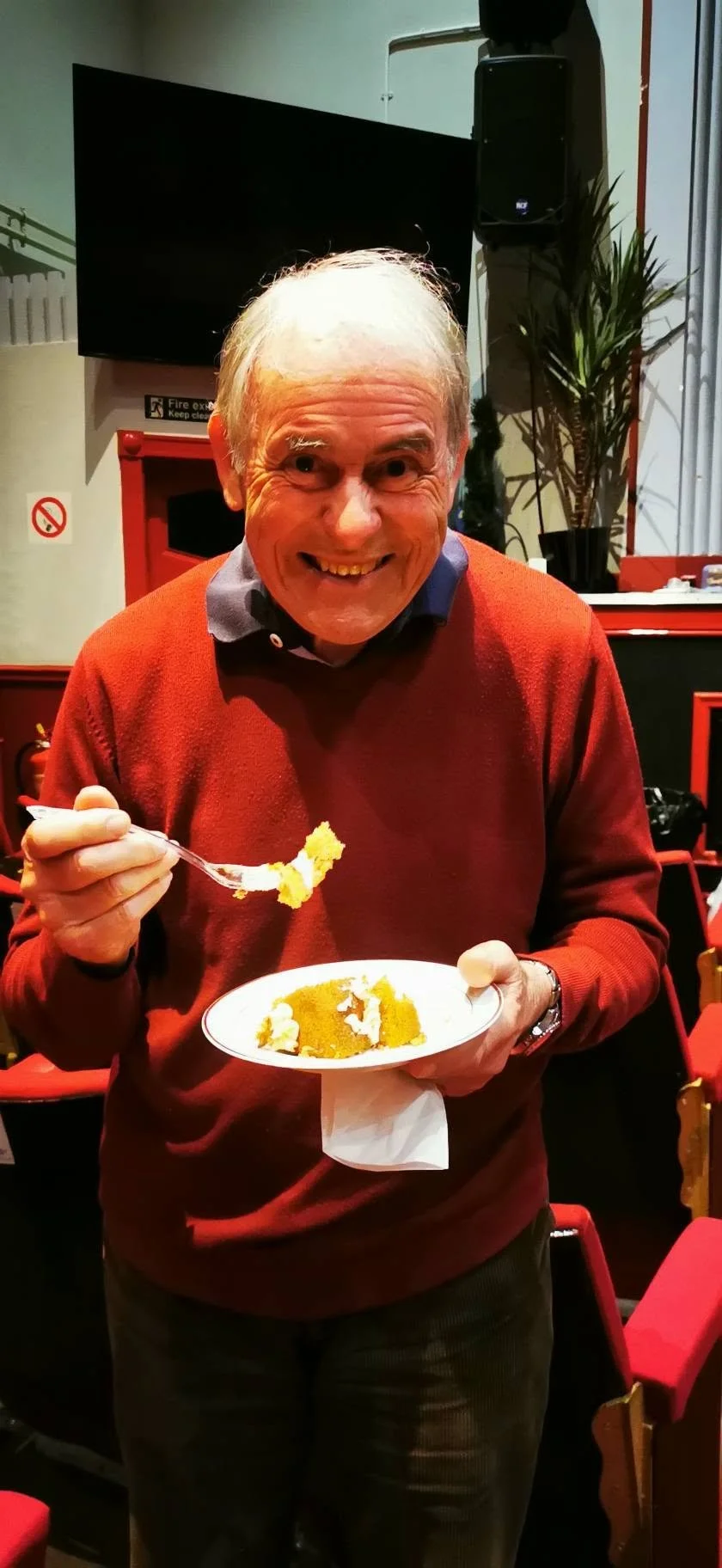 A smiling elderly man in a red sweater holding a plate of yellow cake and a fork, in a restaurant setting.