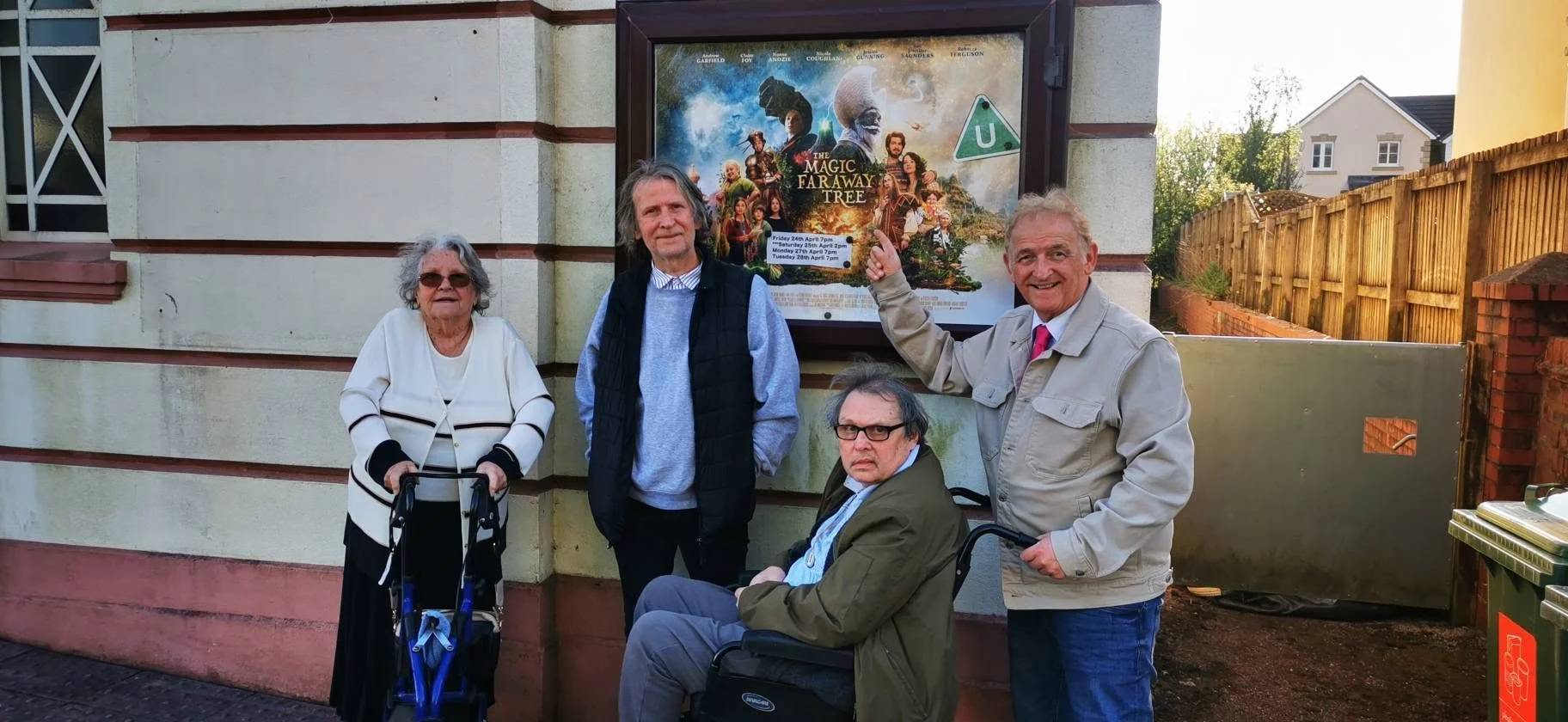 Four people standing outside a building next to a poster for the play 'The Magic Faraway Tree'. One woman with gray hair and sunglasses, using a walker. Two men, one with gray hair and the other with dark hair and glasses, one standing and the other 