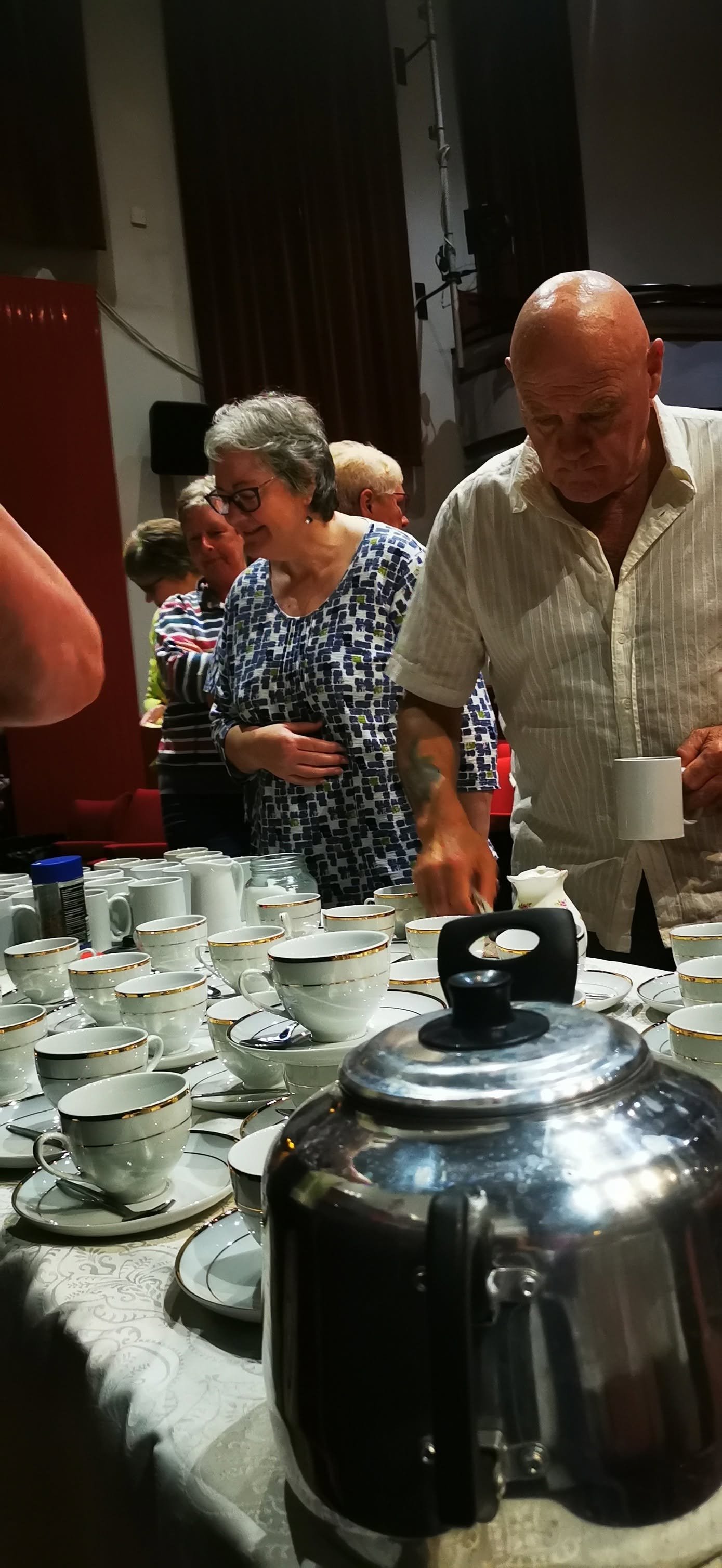 People gathering around a table filled with tea cups and a silver teapot at a social event or tea party.