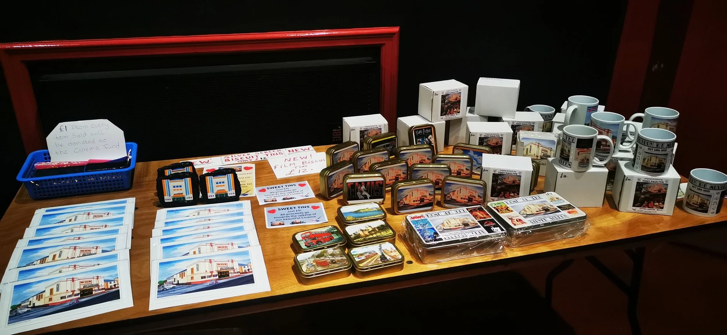 Table displaying various souvenirs including postcards, tins, mugs, and keychains, with signs indicating prices and donation information, against a black background.