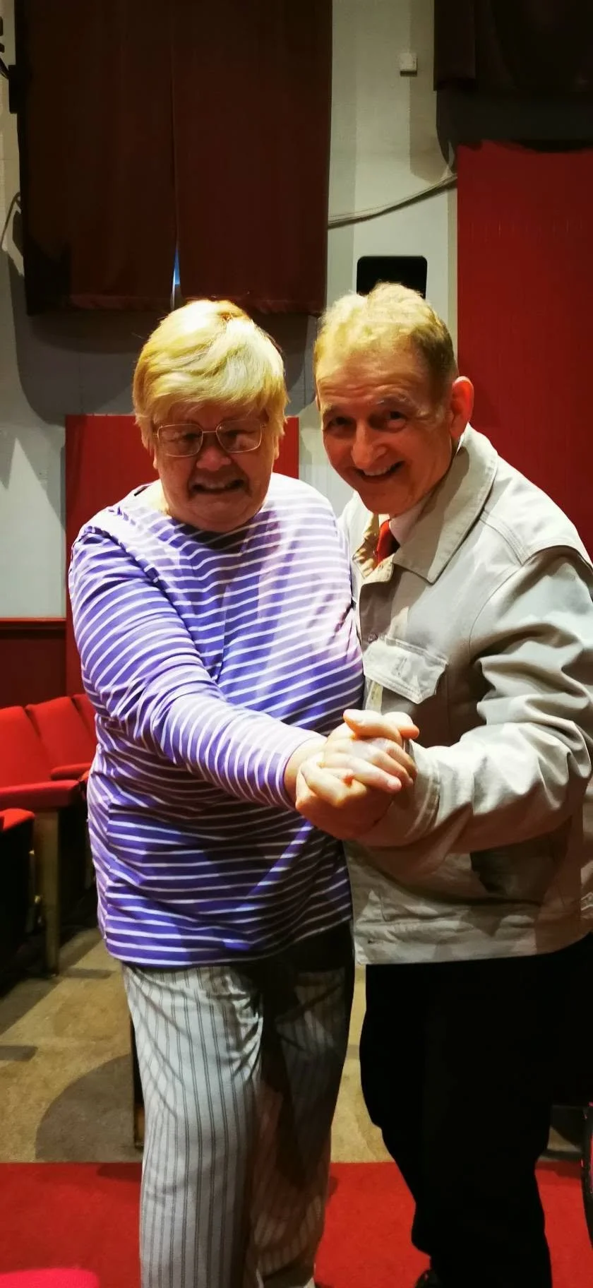 Two elderly men dancing together in a room with red chairs and wall panels, smiling and holding hands.