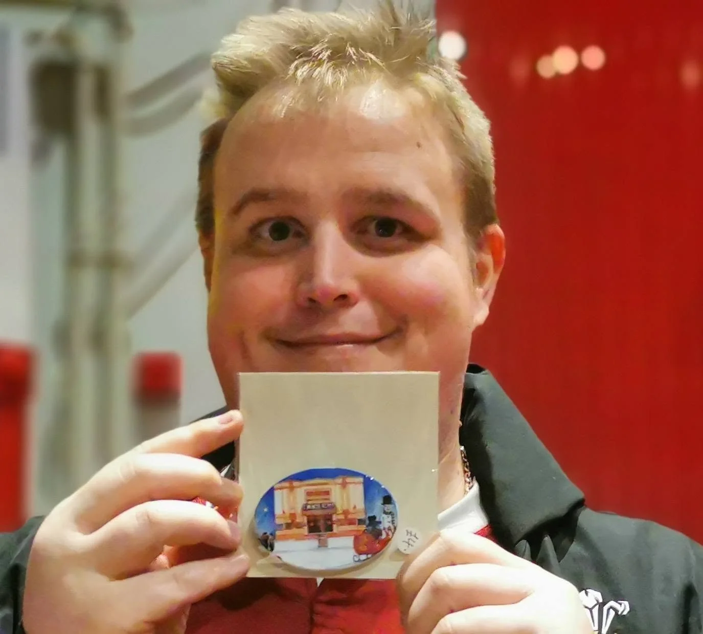 A person with short, light-colored hair smiling and holding a small paper with a sticker of a building in front of their face, inside a room with a red wall in the background.