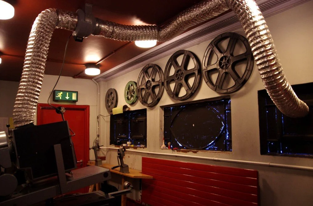 Room with movie projection equipment, vintage film reels on the wall, and a large film reel displayed in the window. Industrial ductwork runs across the ceiling, and there is a red door and illuminated exit sign.