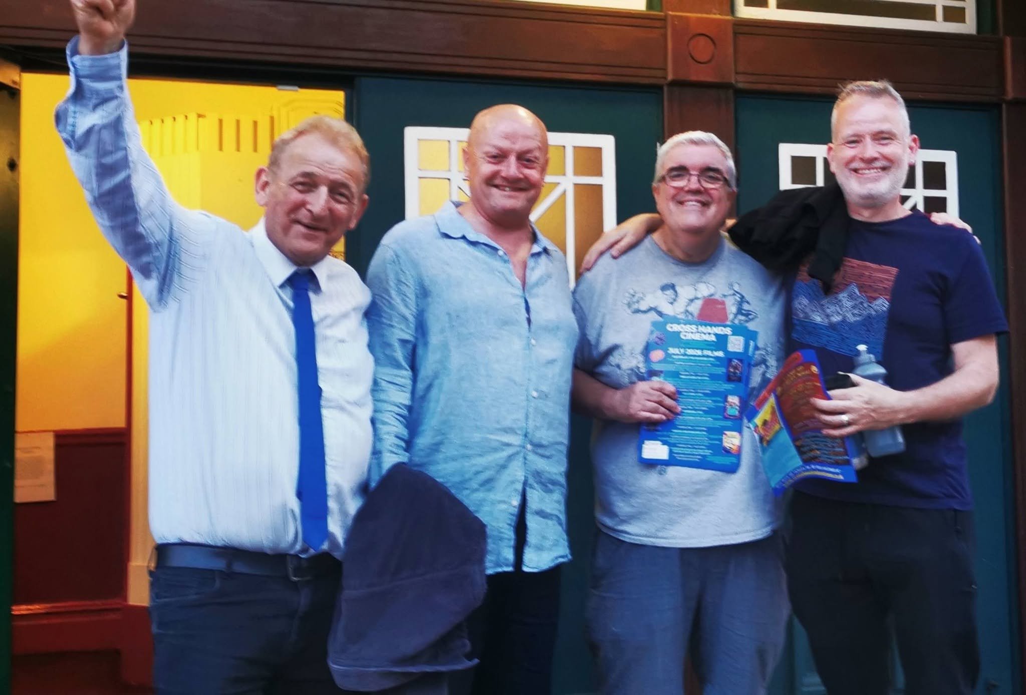 Four men standing together, smiling, in front of colorful background with windows and wooden decor, one man wearing a tie and the others casual clothing, holding brochures, with two men having their arms around each other.