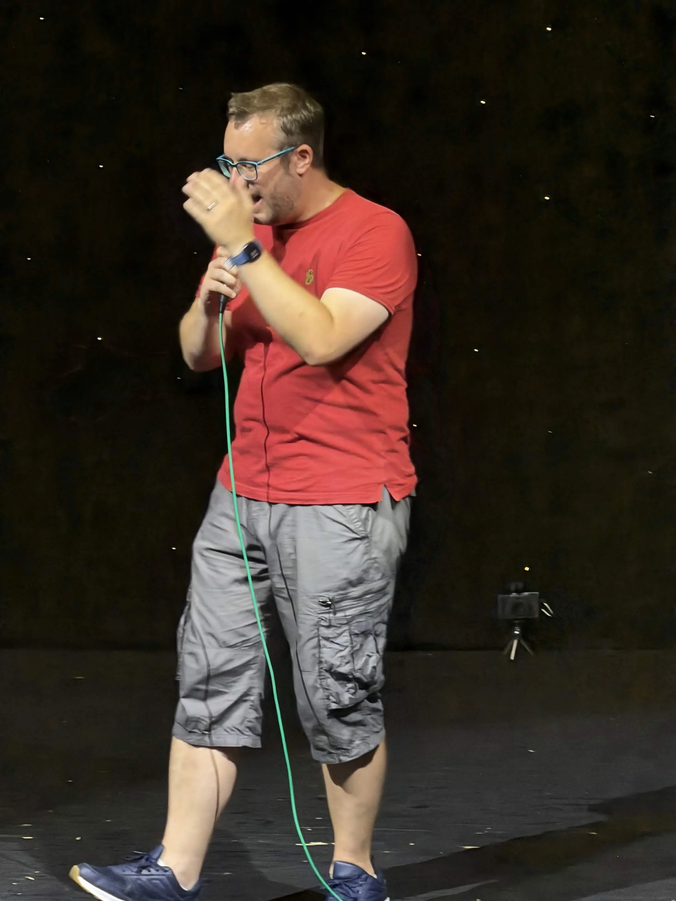 A man wearing glasses, a red shirt, and cargo shorts is on a stage, holding a microphone, and appearing to speak or sing."}