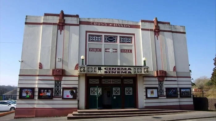 Front view of a historic theater building called Cross-Hands Public Hall with an art deco architectural style, featuring a marquee for Cinema Sensation and four posters outside.