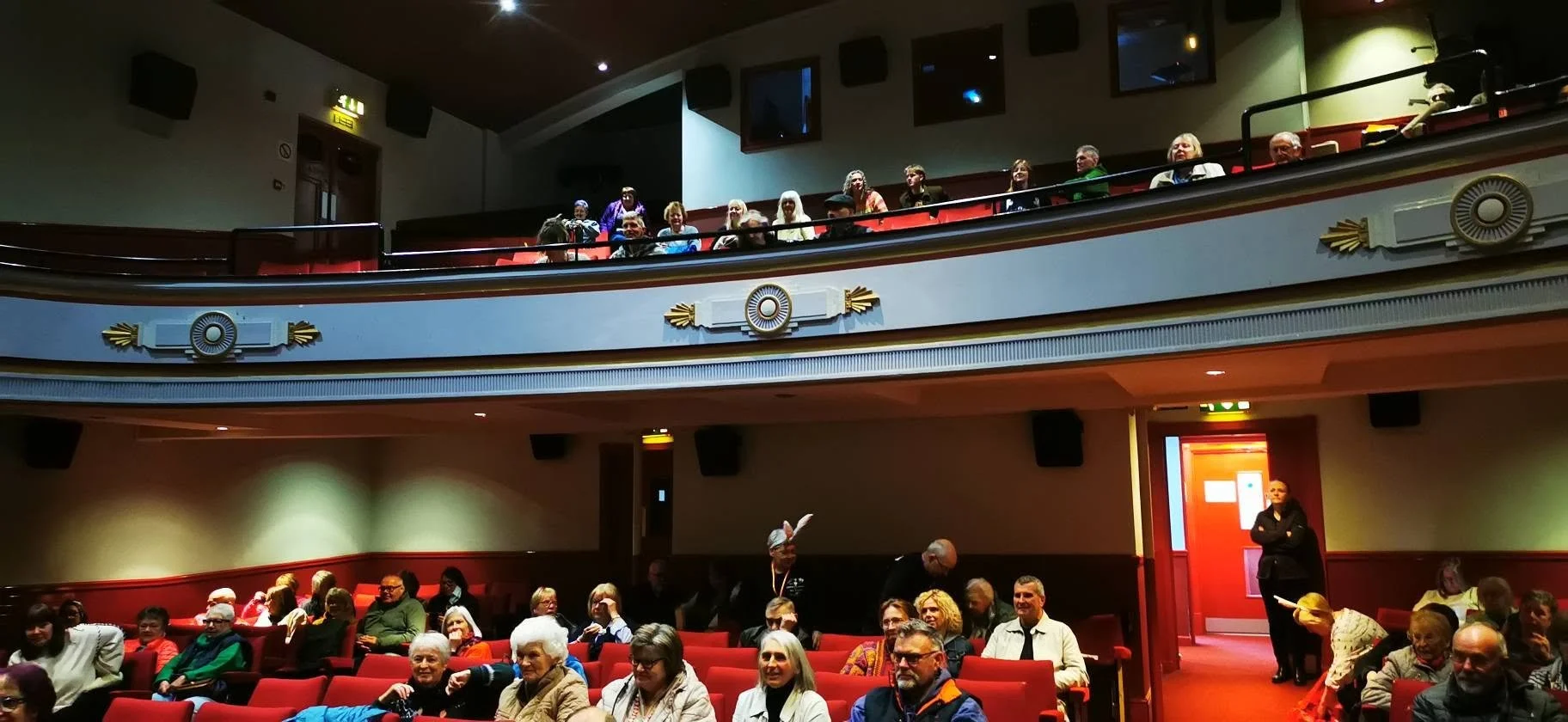 Audience in a theater or auditorium with red seats and a balcony, some people are standing and others sitting, ready for a performance or event.