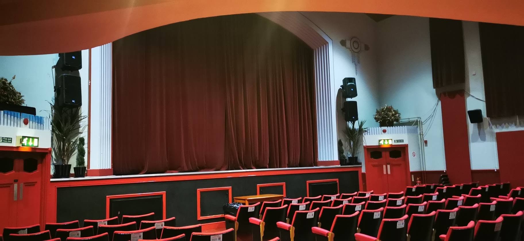Empty theater auditorium with a stage, closed brown velvet curtains, red and black seating, and potted plants near the stage.