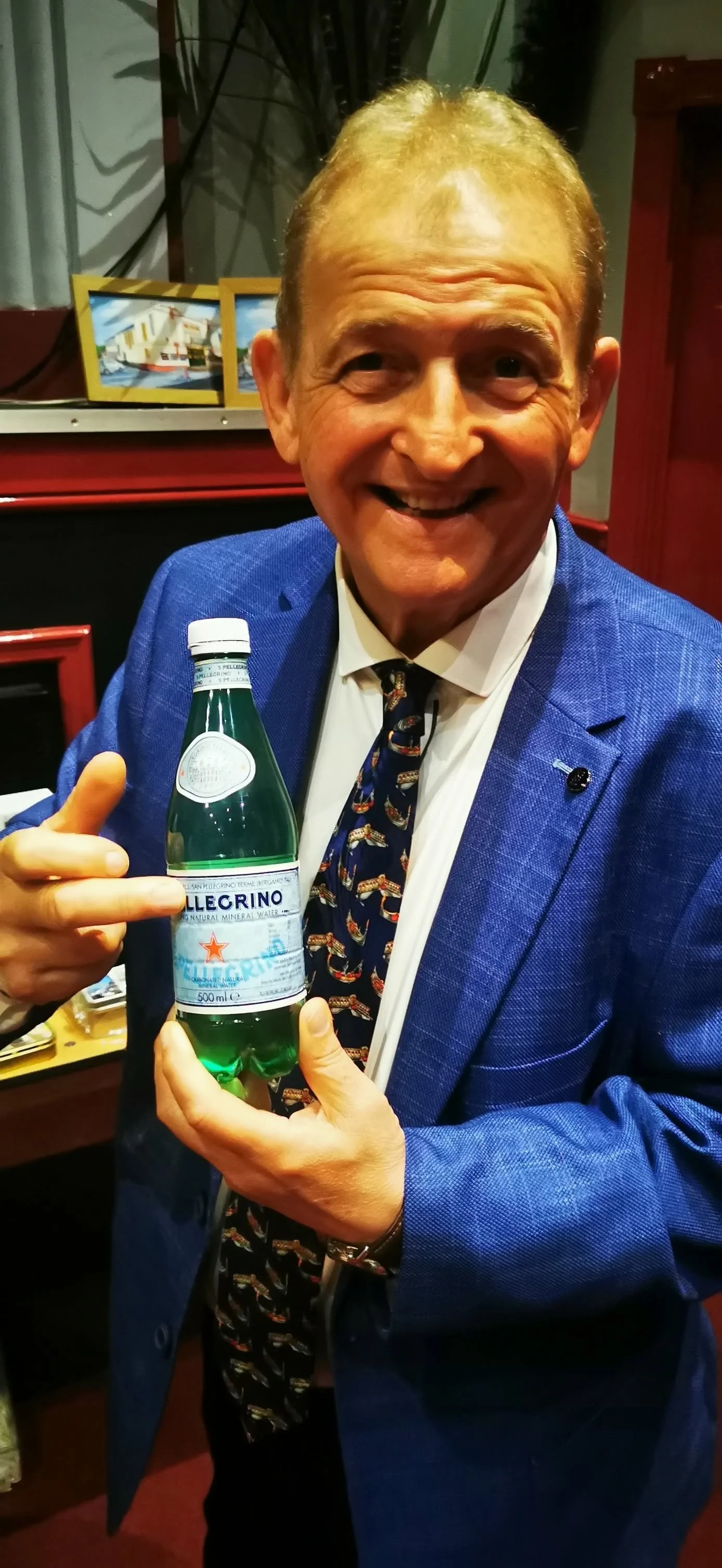An older man with short, blond hair smiling and pointing at a bottle of San Pellegrino sparkling mineral water he is holding. He is wearing a blue suit jacket, a white shirt, and a navy tie with a printed design. The background includes framed pictur
