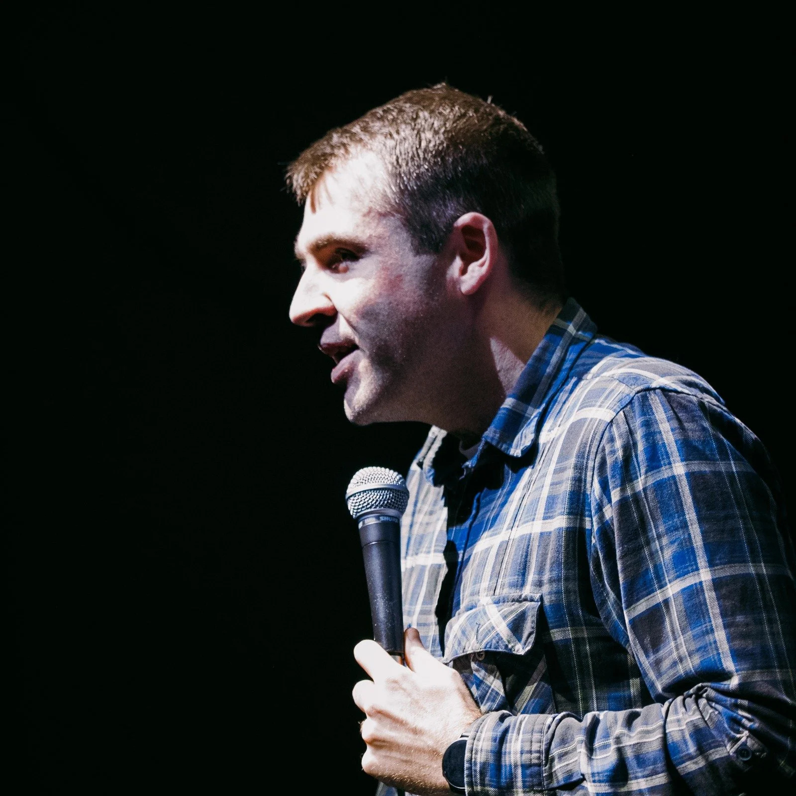 A man holding a microphone, wearing a blue and black plaid shirt, standing against a dark background.