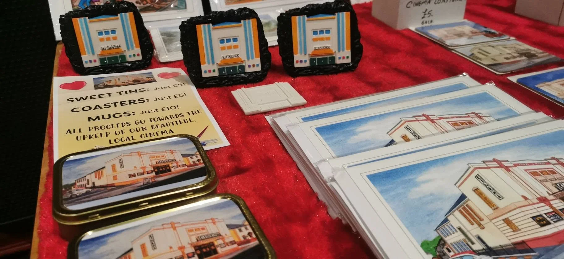 Decorative magnets, postcards, and promotional flyers on a red tablecloth at a market stall. The magnets depict a building facade, and the flyers promote local cinema fundraising.