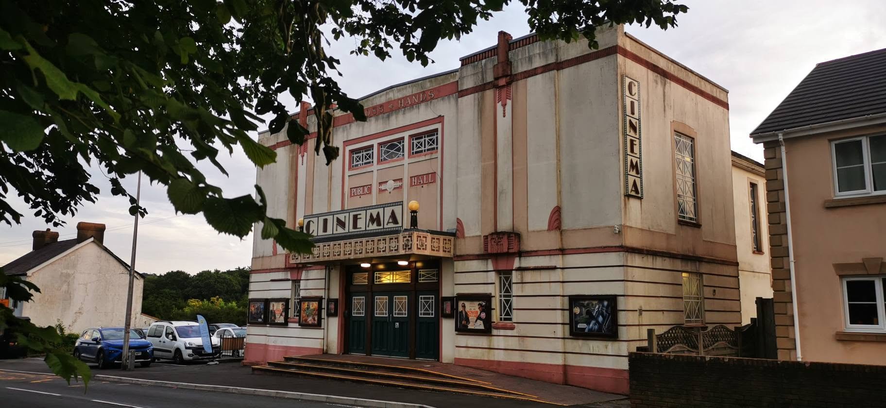 The image shows an old building with a vintage cinema sign on it. The building's architecture is classic, with decorative elements and poster displays outside the entrance. There are parked cars and trees partially framing the shot, and the scene app