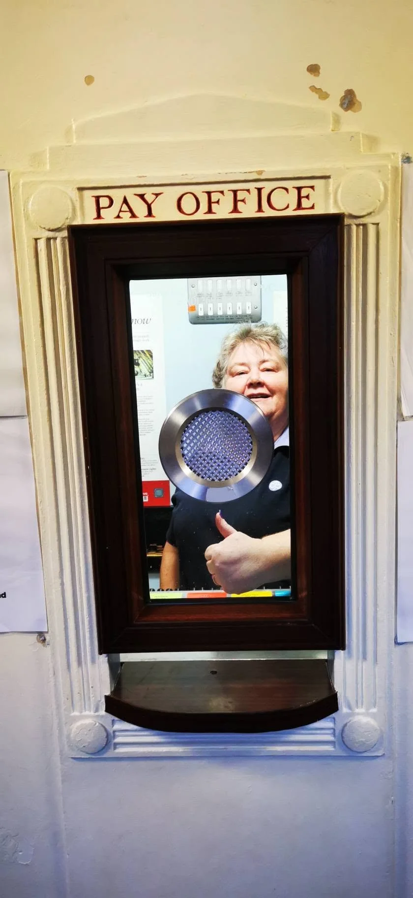 A woman smiling and giving a thumbs-up through a window at a pay office, with a metal plate in front of her face.