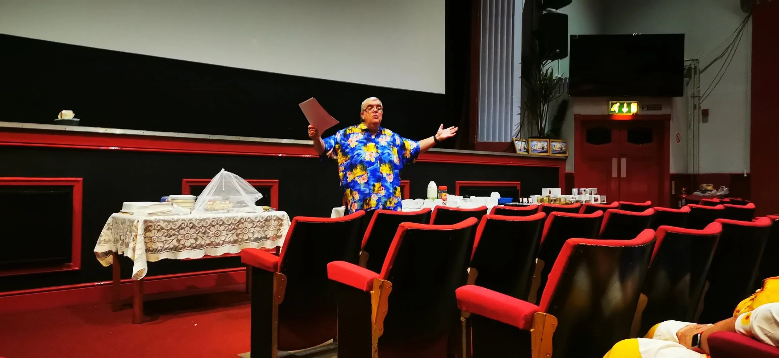 A man wearing a colorful Hawaiian shirt stands in front of an empty auditorium or theater, gesturing with his arms open. There is a table with covered food items and drinks in front of him, surrounded by red theater seats.