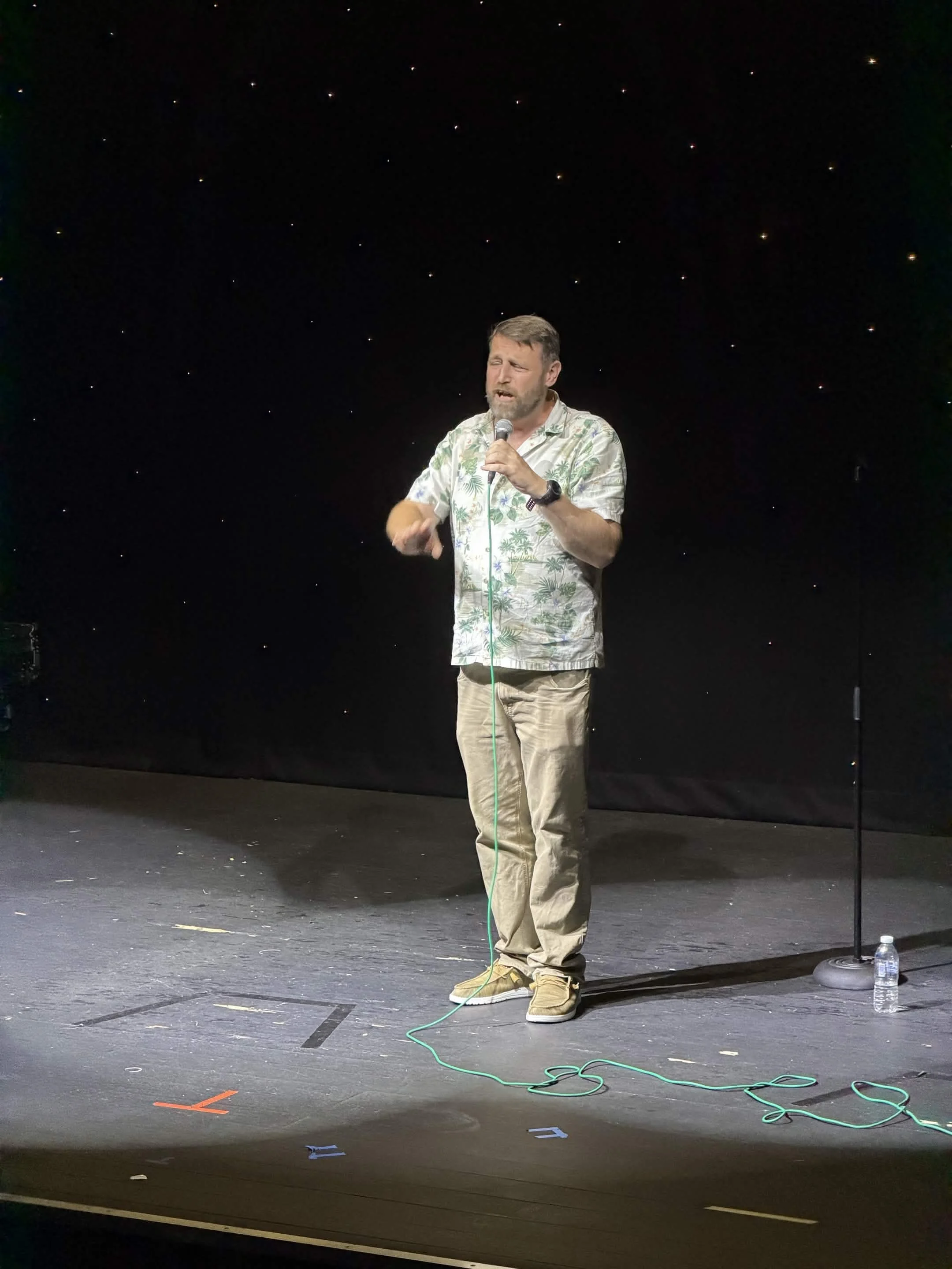 A man with a beard and short hair, wearing a short-sleeved Hawaiian shirt and beige pants, is speaking or singing into a microphone on a stage with a black starry background. There is a water bottle on the floor to his right, and colorful tape markin