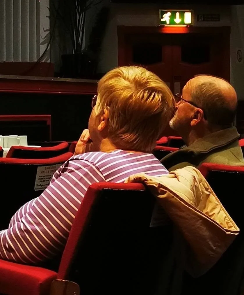 A woman and a man sharing a kiss in a theater or auditorium with red seats, an emergency exit sign glowing green above them, and a potted plant in the background.