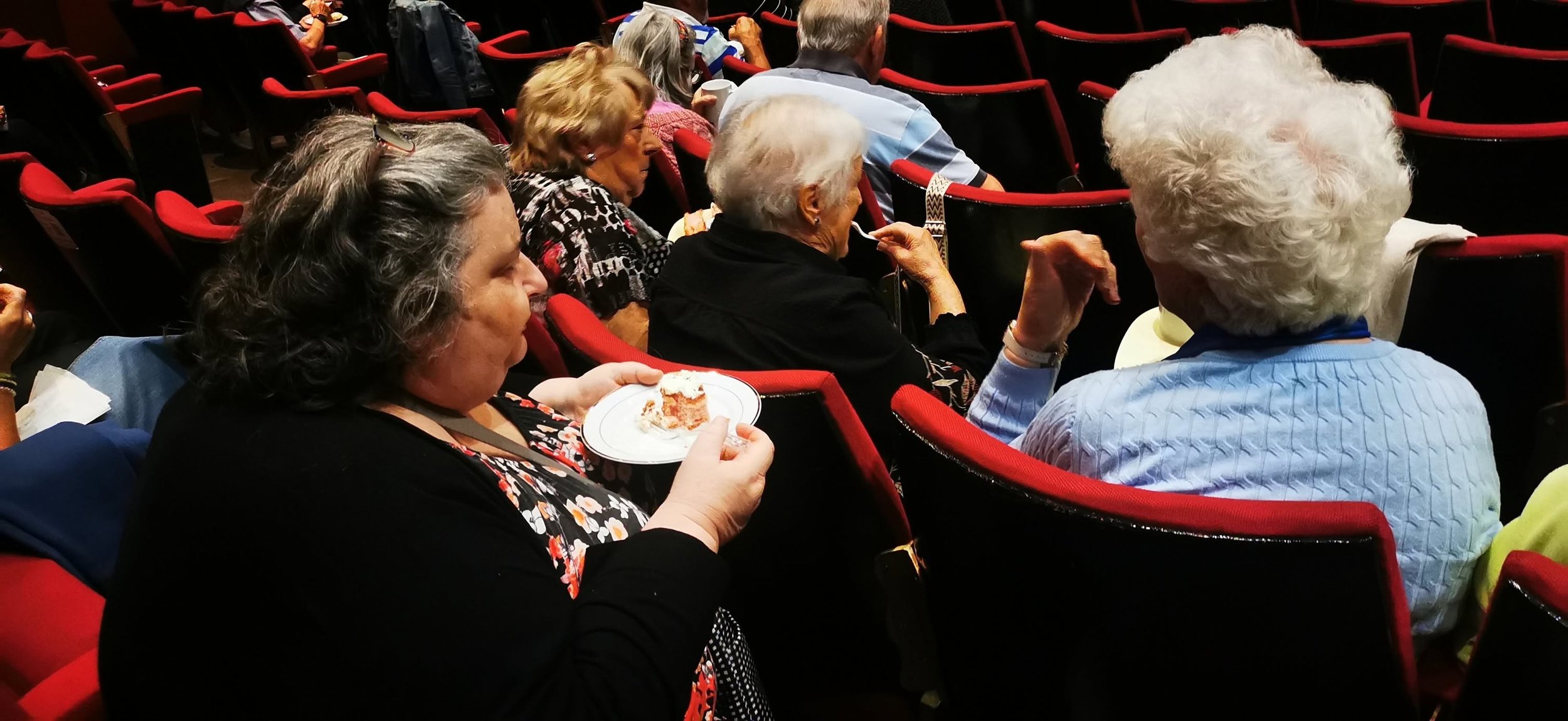 Group of elderly people sitting in theater seats, some eating cake, engaged in conversation, with mostly empty red and black theater seats around them.