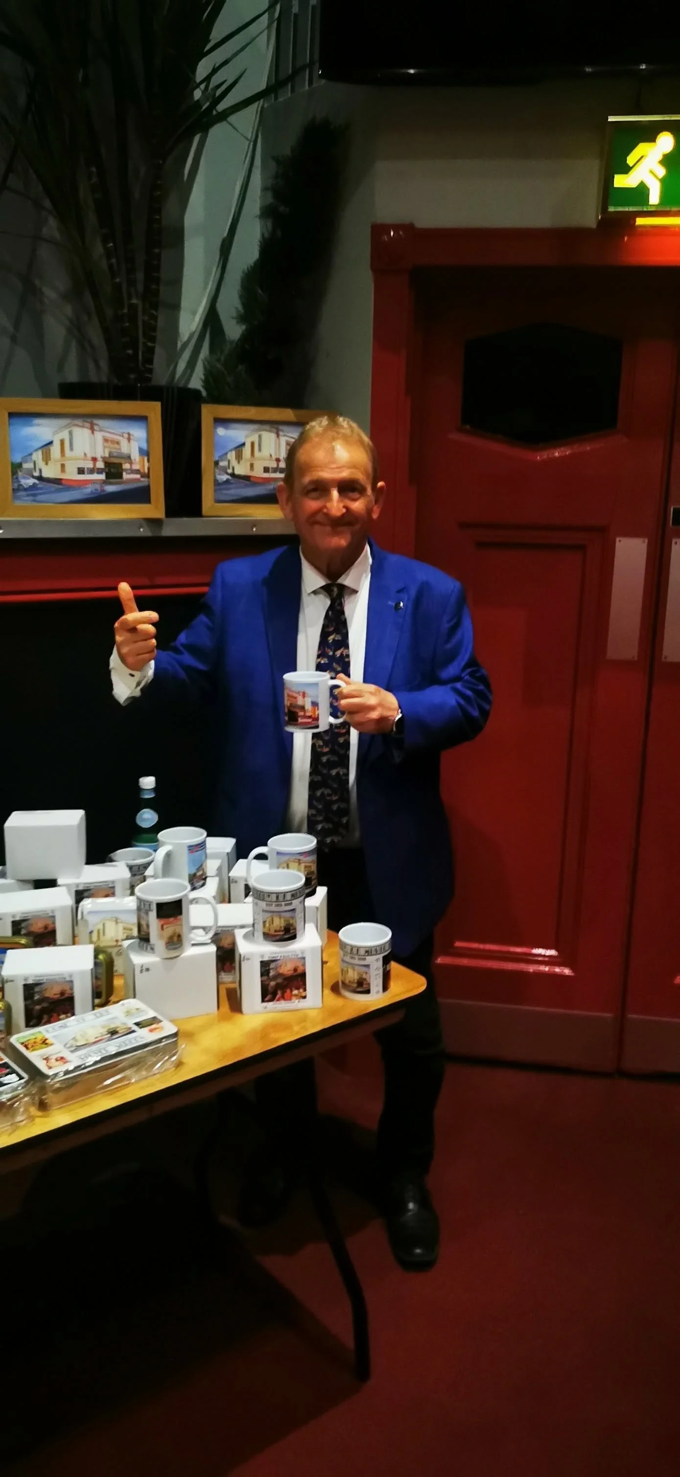 A man in a blue suit holding a mug stands behind a table with multiple mugs and other souvenir items featuring an illustration of a building. The man is smiling and giving a thumbs-up. There are pictures of the building on the wall behind him, and a 