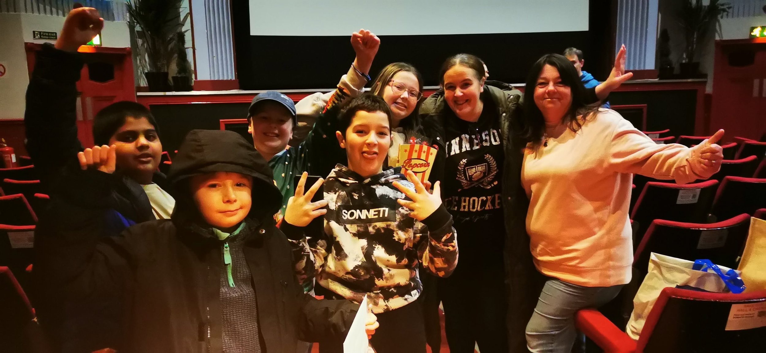 Group of children and women posing together in a theater or auditorium with red chairs and a large screen in the background. The children are smiling and making hand gestures; some are wearing jackets and hoodies.