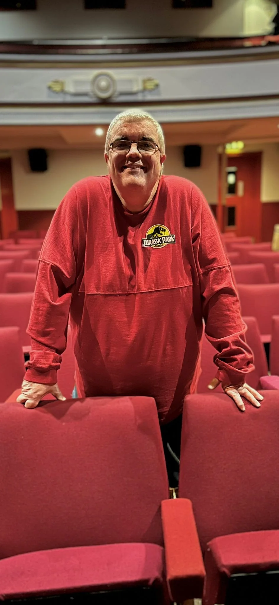 A man standing behind theater seats, wearing a red hoodie with a Jurassic Park logo. The theater is empty with red seats and a prominent ceiling decoration.