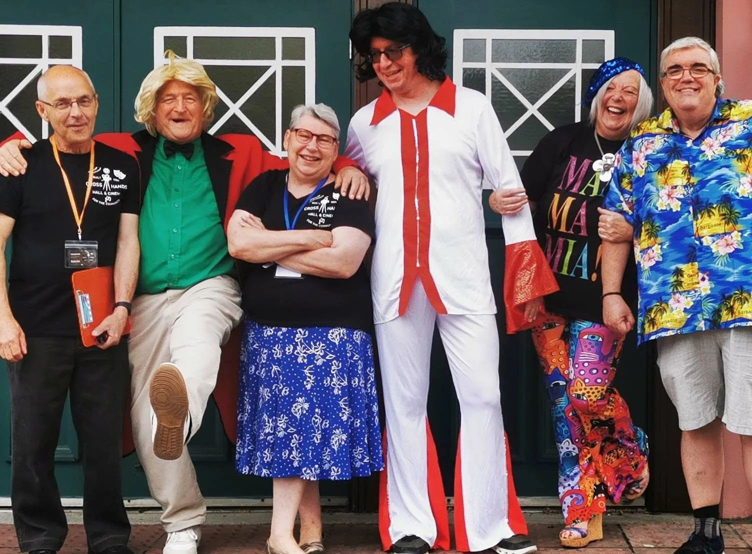 A group of six people dressed in colorful and retro outfits, standing in front of a green door, smiling and posing for the photo.