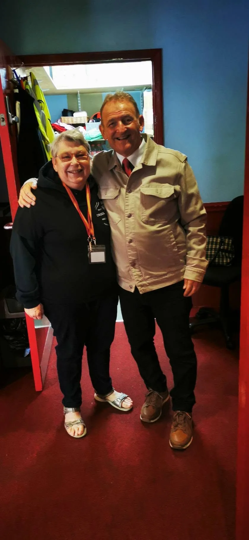 Two smiling people, a woman on the left and a man on the right, standing indoors on a red carpet, with the woman wearing a black hoodie and sandals, and the man wearing a beige jacket and brown shoes, in front of a blue wall and a doorway with shelve
