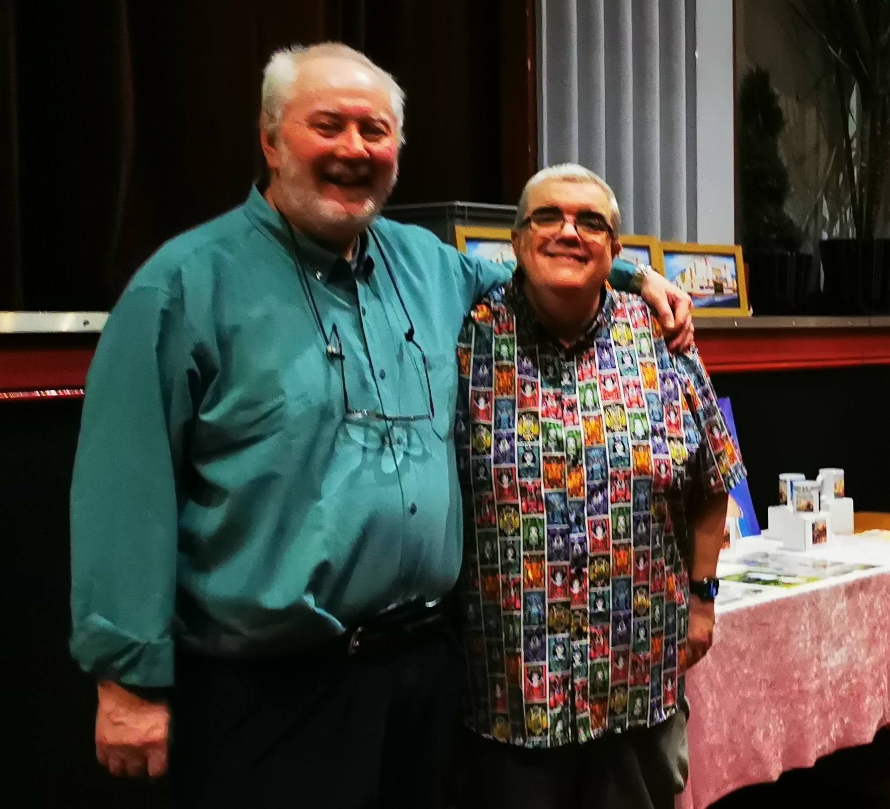 Two smiling men standing side by side at an indoor event, one wearing a teal shirt with glasses around his neck, the other wearing a colorful, patterned shirt with a collection of small framed art pieces in the background.