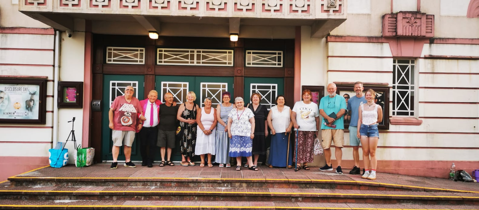 A group of fifteen people standing on steps outside a building with green doors and pink architectural details, posing for a photo.