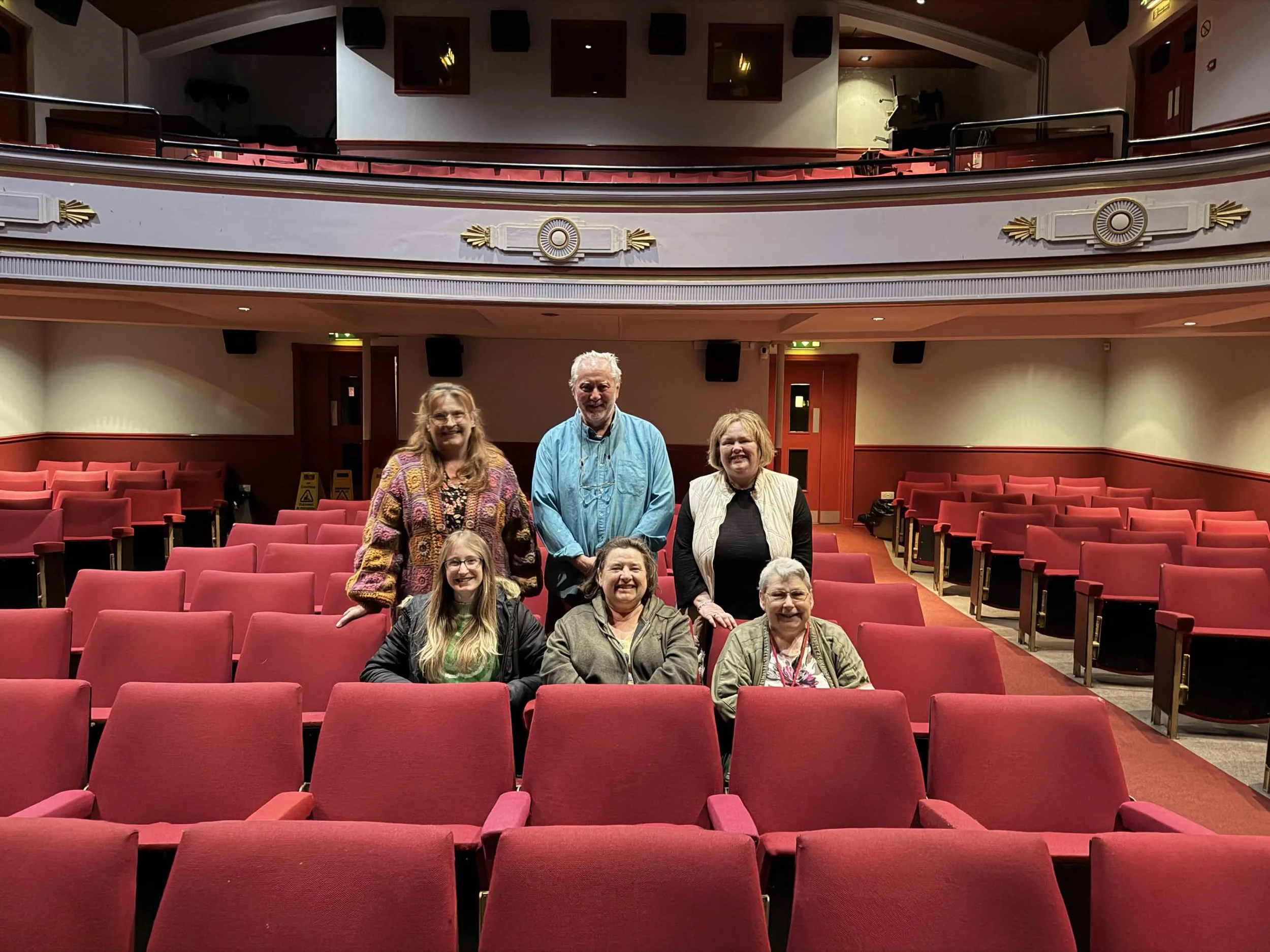 A group of nine people standing in an auditorium with red seats and decorative gold and white trim. The auditorium has a balcony area with red seats and black speakers mounted on the wall.