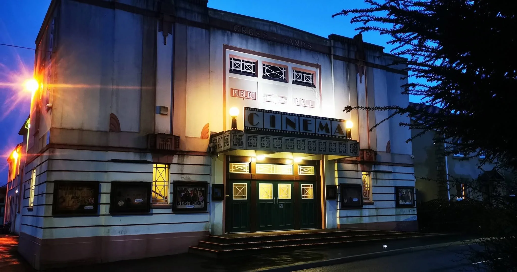 A vintage-style cinema building with a marquee that reads 'CINEMA,' illuminated by exterior lights, with posters displayed in the windows, and a small balcony above the entrance, set against a dark blue evening sky.