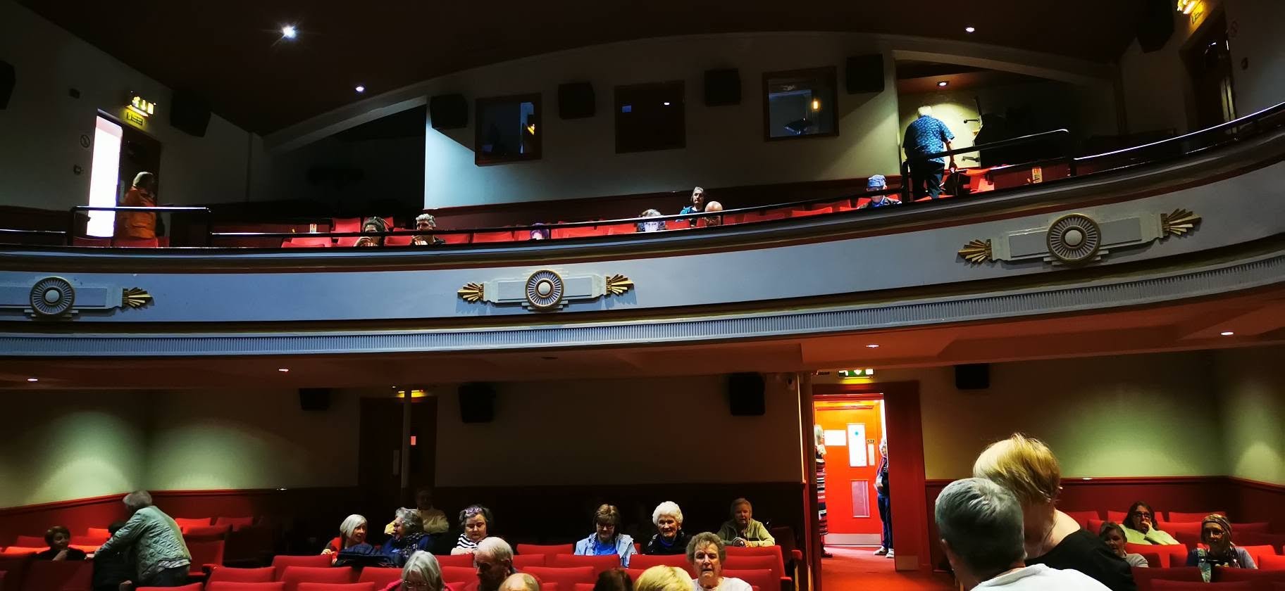 People sitting in red theater seats inside a dimly lit auditorium, with some standing and talking near a door, and others on the balcony level.