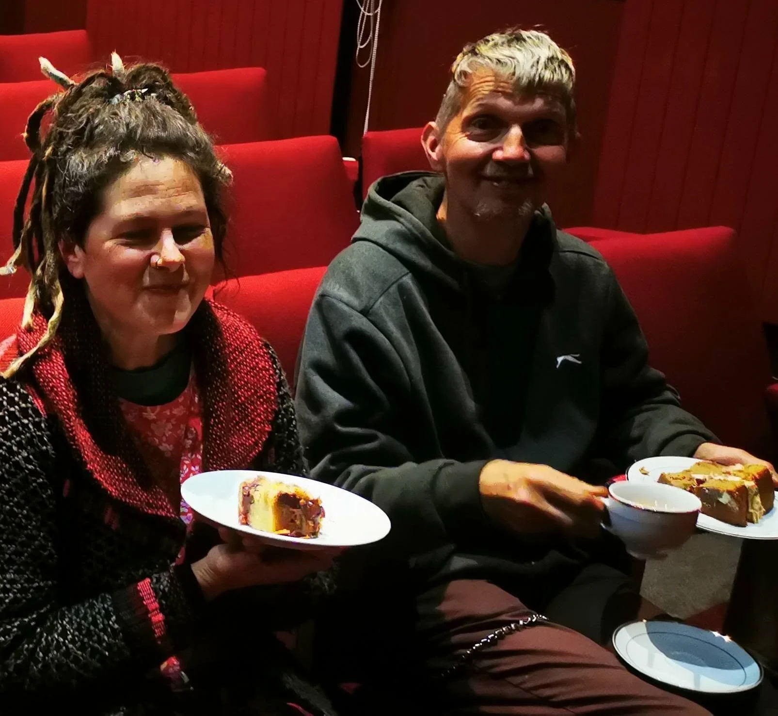 Two people sitting in a theater holding plates with slices of cake and bowls of dessert, smiling at the camera. The woman has dreadlocks and is wearing a red and black patterned top, while the man has short gray hair and is dressed in a black hoodie.