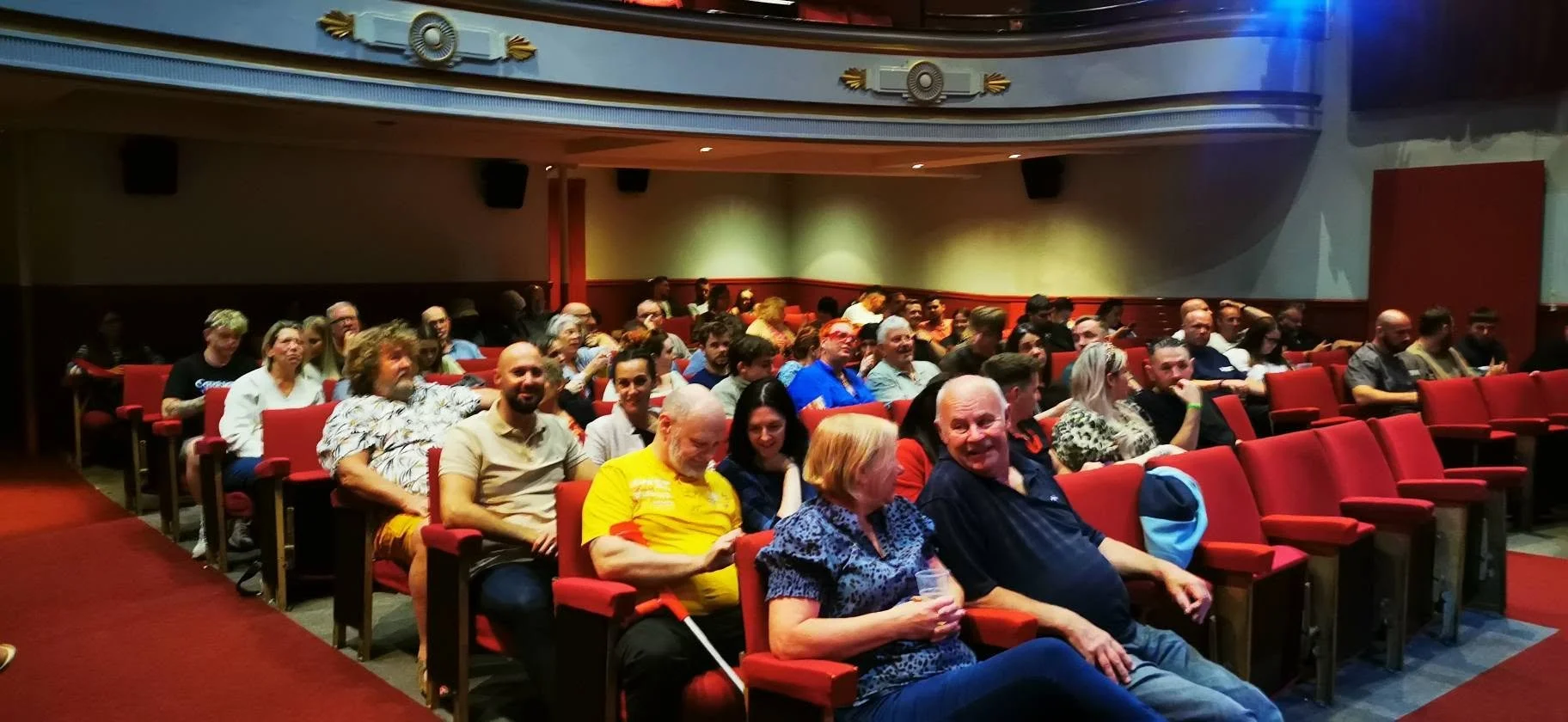 Audience seated in a theater, some smiling and engaging, with red theater seats and a large blank screen or wall to the right.