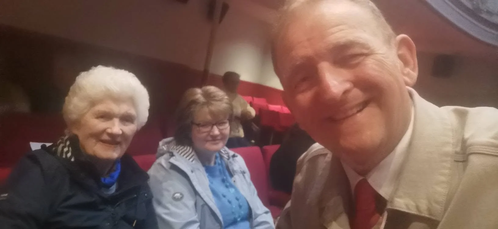 A man taking a selfie with two elderly women sitting in theater seats.