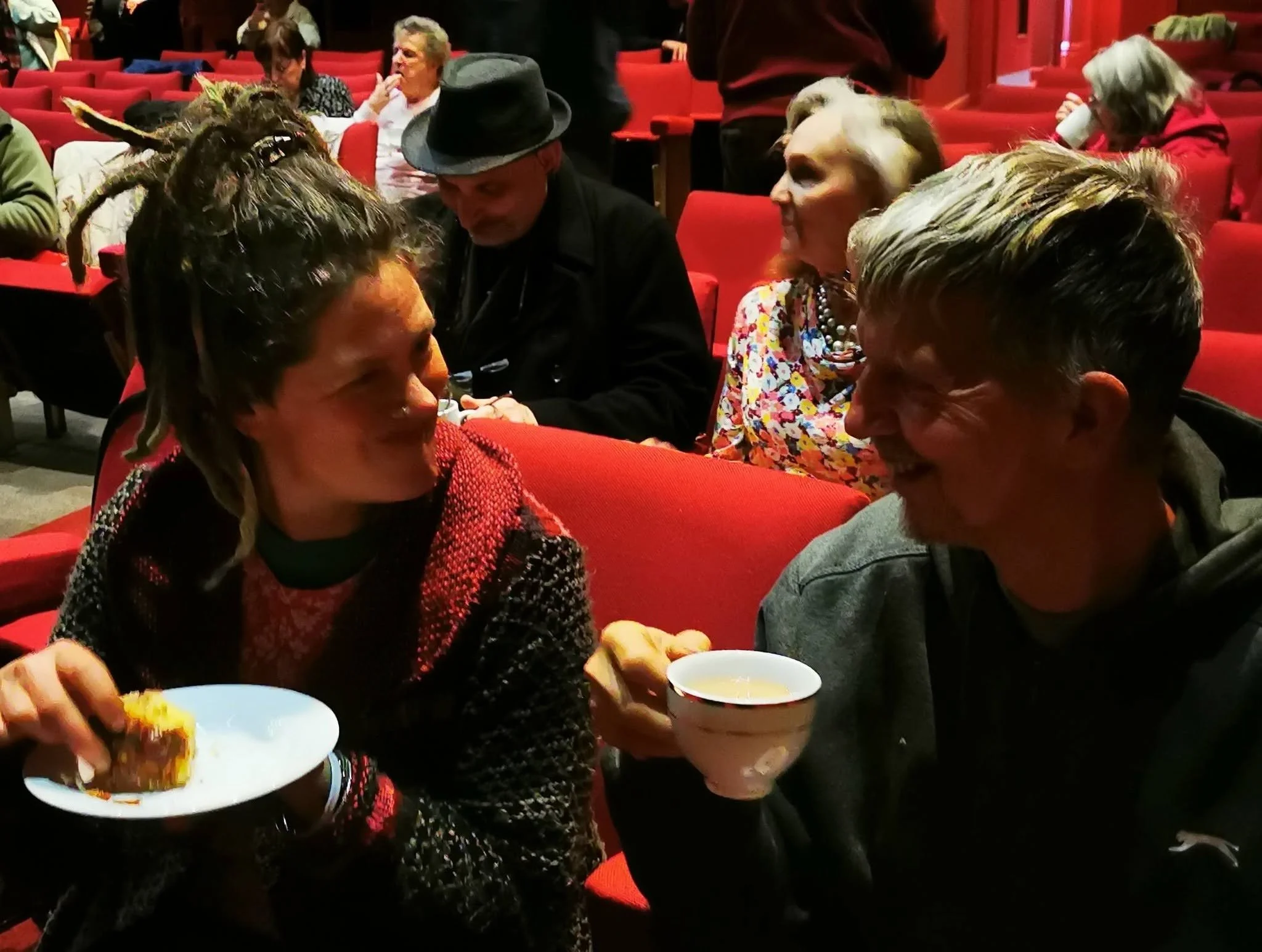 People sitting in red theater seats, eating and drinking, some wearing costumes or distinctive clothing, in a dimly lit setting.