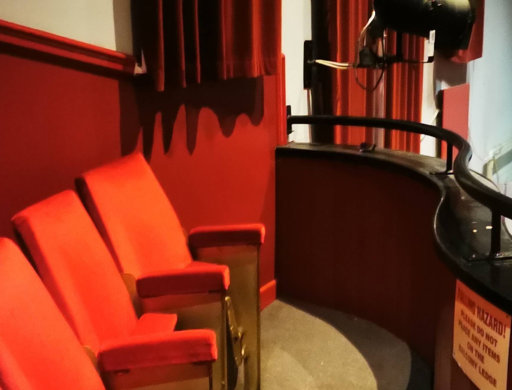 Empty red theater seats with armrests, facing a red curtain on the wall behind them, in a dark room.