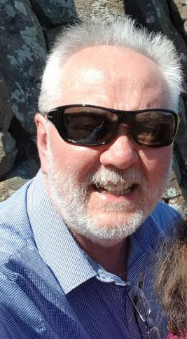 Close-up of a smiling older man with white hair and beard, wearing sunglasses and a blue shirt, outdoors with rocks in the background.