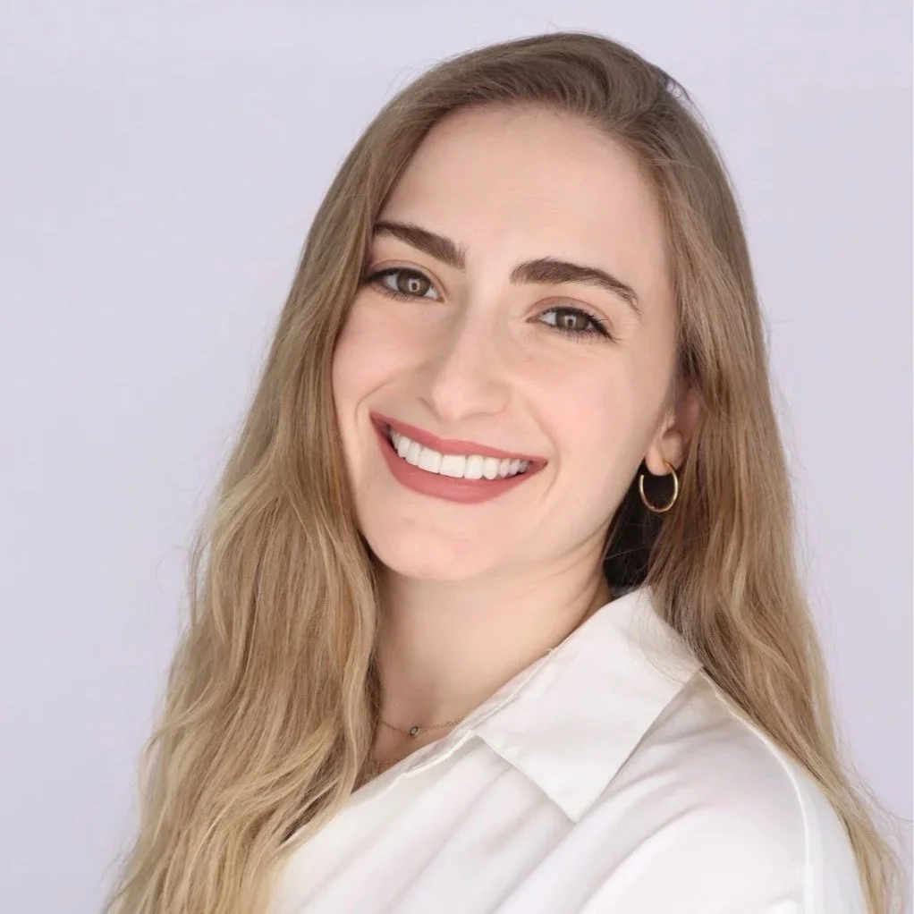 A young woman with long blonde hair, smiling, wearing a white shirt, gold hoop earrings, and a small necklace, against a light gray background.