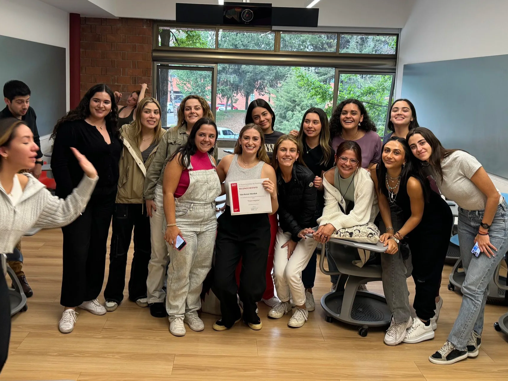 Group of diverse young women and men in a classroom celebrating, with one woman holding a certificate and smiling at the camera.