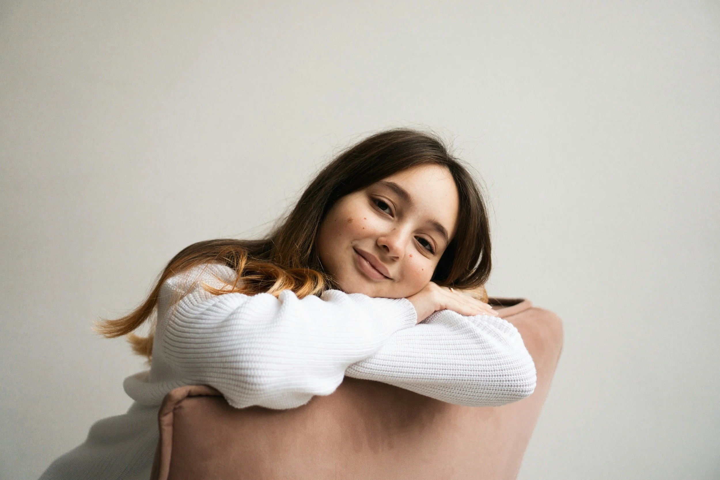 A young girl with light brown hair and a smile, resting her head on her crossed arms, being embraced by an adult.
