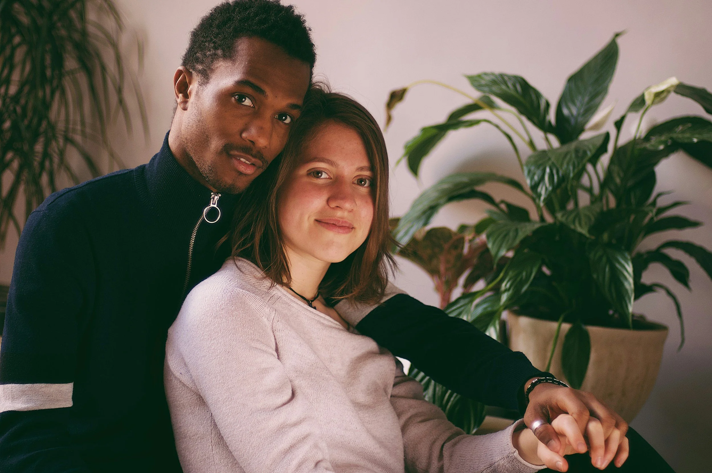 A Black man and a white woman sitting close together inside, with a large green houseplant behind them. The man is wearing a dark zip-up sweater, and the woman is wearing a pink sweater. They are both smiling softly.