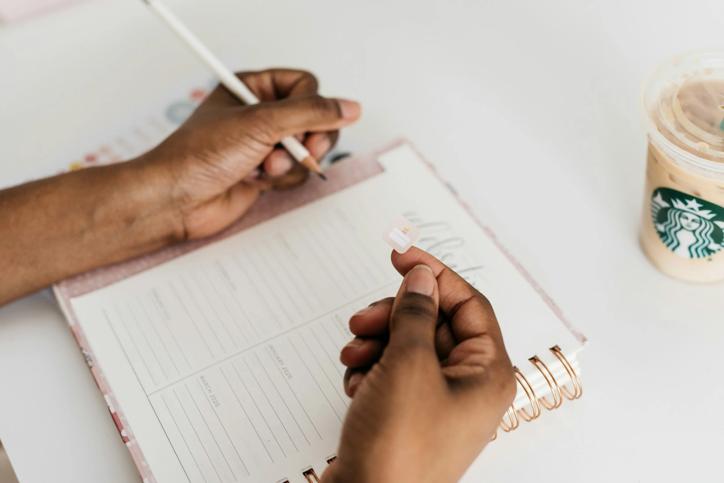 A person holding a small SIM card between their fingers while writing in a planner with a pen. A Starbucks iced coffee drink is on the right side of the image.