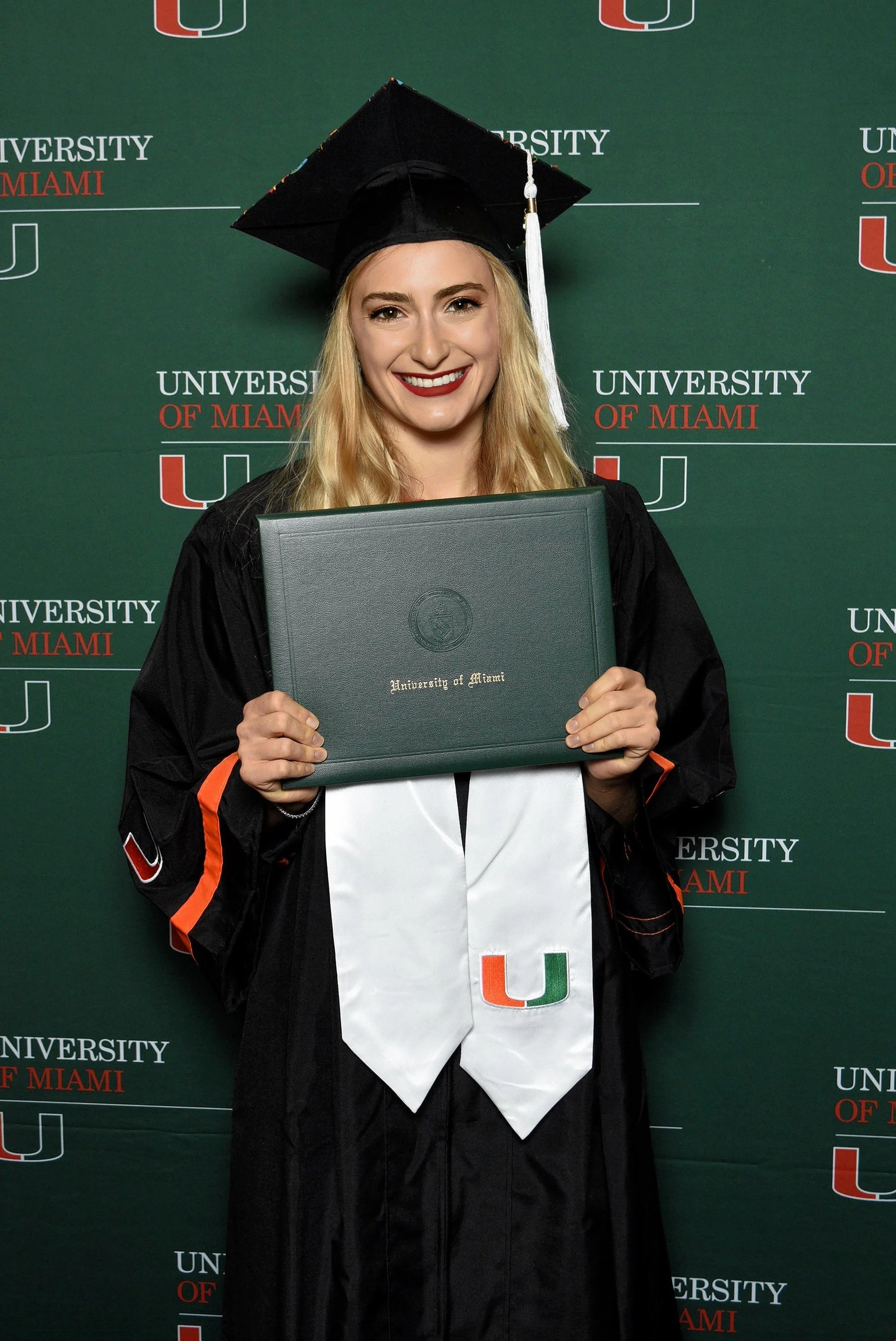 A young woman in a graduation cap and gown holding a diploma in front of a backdrop with the University of Miami logo.