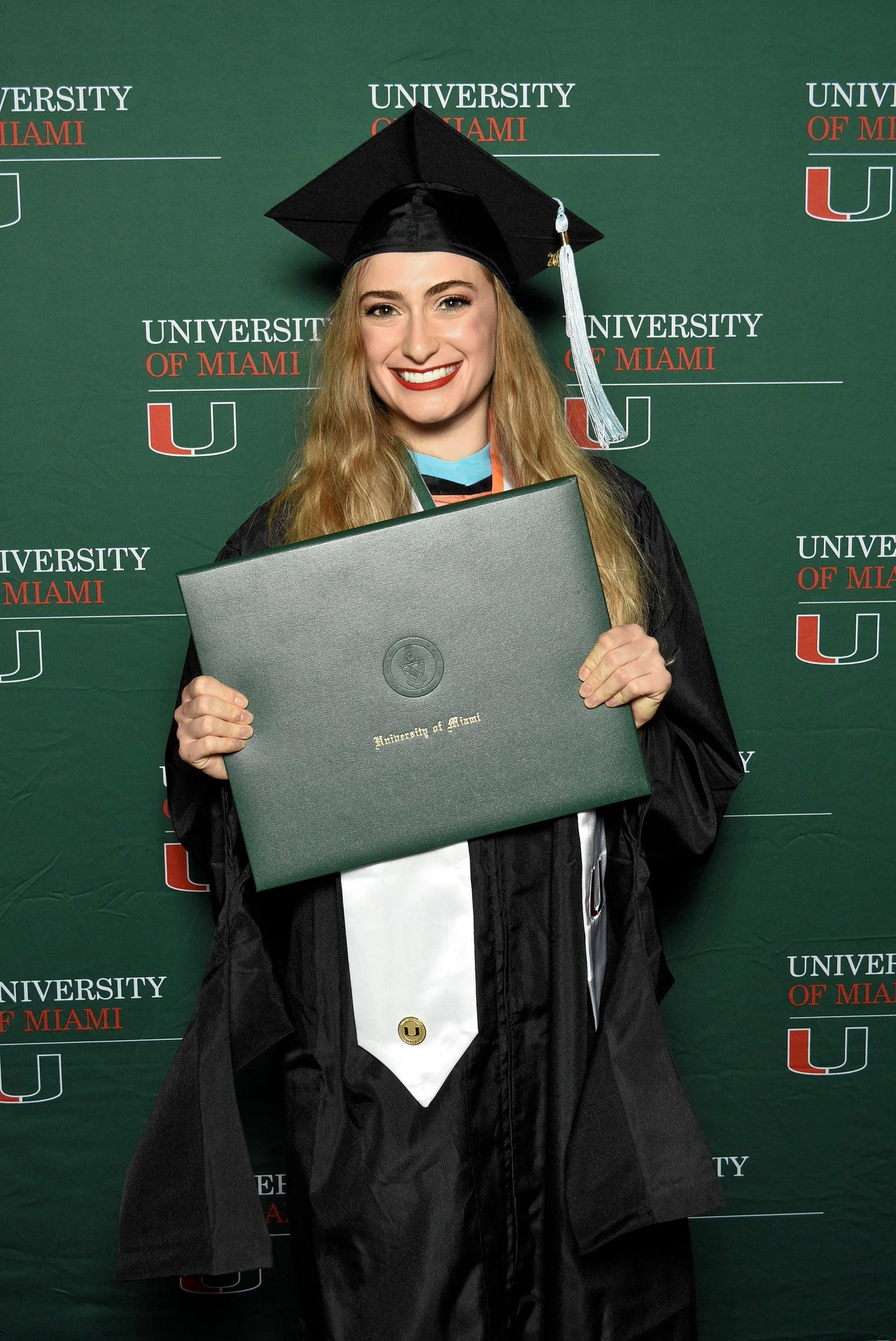 A woman in graduation attire holding a diploma in front of a University of Miami backdrop.