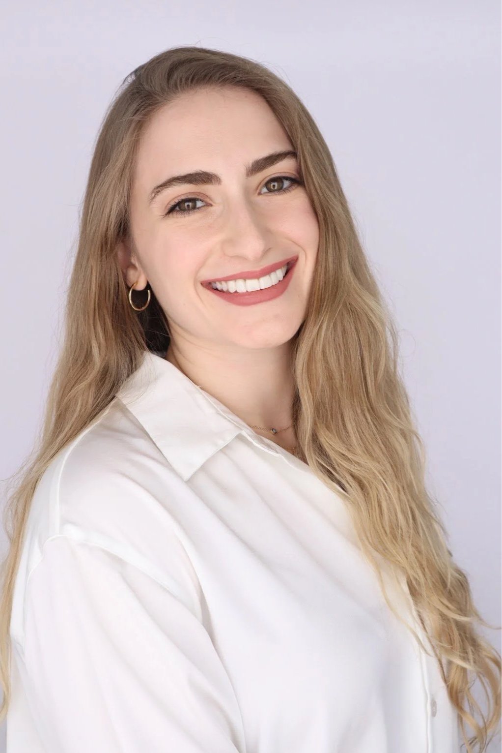 A young woman with long, wavy blonde hair, wearing a white button-up shirt, hoop earrings, and a subtle necklace, smiling against a plain light background.