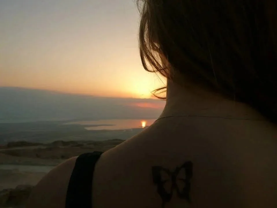 Woman with brown hair viewing sunset over a body of water, wearing a black strap top, with a butterfly tattoo on her shoulder.