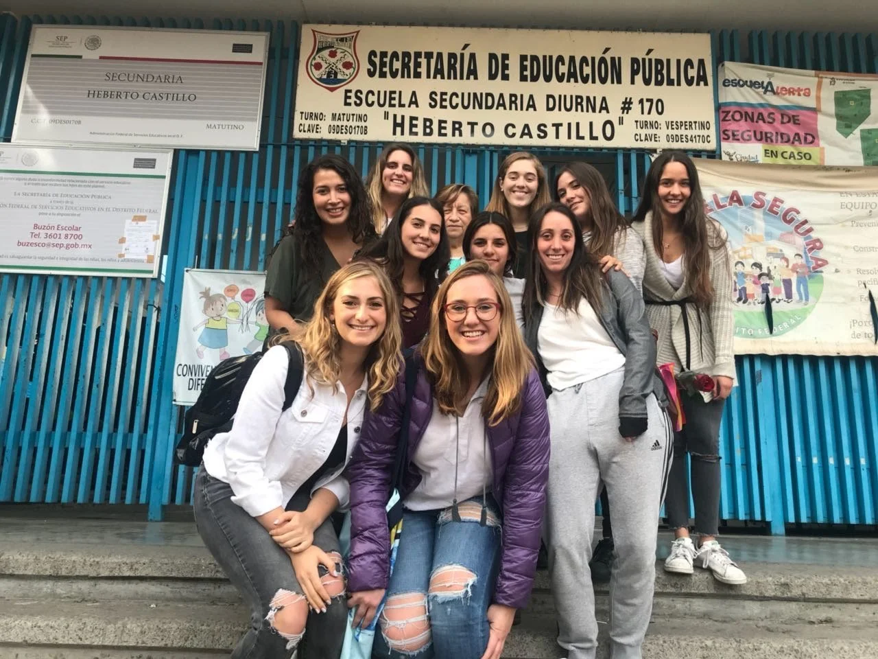 Group of teenage girls smiling and posing for a photo outside a school building with signs and posters in Spanish.