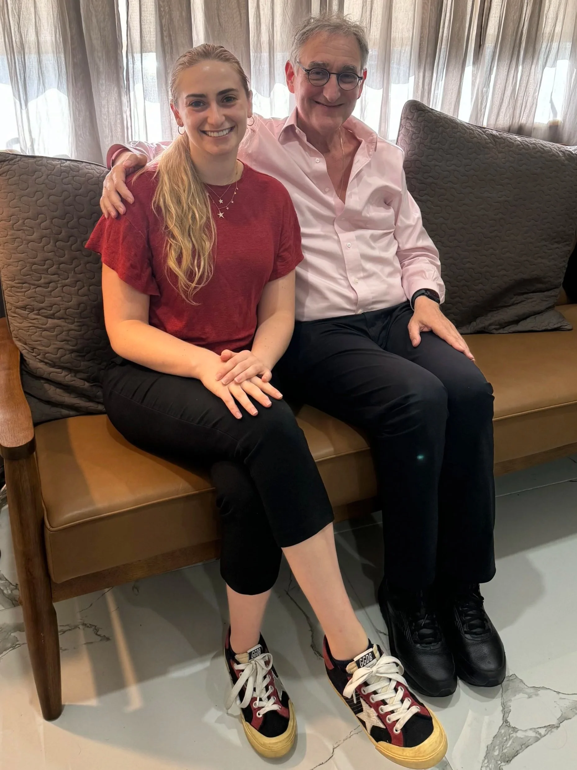 A young woman with long blonde hair in a ponytail, wearing a red T-shirt, black pants, and colorful sneakers, sitting next to an older man with glasses, wearing a pink shirt and black pants, on a wooden bench with large gray pillows, in front of beige curtains.